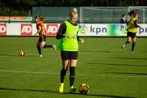 Eine Gruppe von Frauen, die auf einem von Bäumen umgebenen Feld Füssball spielen, mit Bannern im Hintergrund und einem Netz im Vordergrund.