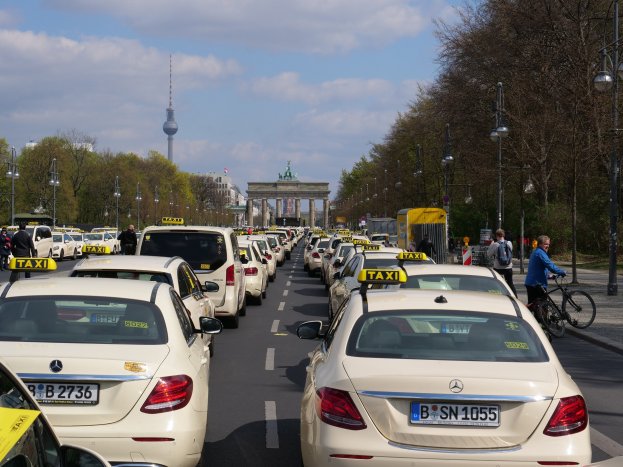 Eine lange Reihe von Taxis, die an einer belebten Straße in Berlin, Deutschland, geparkt sind, mit Fahrradfahrern und Fußgängern auf dem Gehweg, flankiert von Laternen und Bäumen, und Gebäuden, einem Bogen und einem Turm im Hintergrund unter einem bewölkten Himmel.