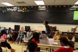 Ein Klassenzimmer mit Kindern an Tischen mit verschiedenen Gegenständen, einer Lehrerin in der Mitte vor einer Tafel und Taschen auf dem Boden.