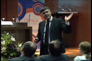 Ein Mann in einem schwarzen Blazer steht an einem Podium mit Mikrofonen und spricht in ein Handmikrofon, während in der Nähe ein Blumentopf oder ein Blumenstrauß steht. Vor ihm sitzen zwei Männer und eine Frau auf Stühlen. Hinter ihm befindet sich ein Tisch mit einem Buch, einer Wasserflasche und anderen Gegenständen, sowie einer braunen Wand und einem bunten Schild mit Text im Hintergrund.