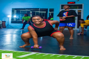 Eine Gruppe von Menschen in einem Fitnessstudio mit einer Frau, die mit Hanteln in der Kniebeugeposition sitzt, umgeben von Geräten und einem Hintergrundbild.