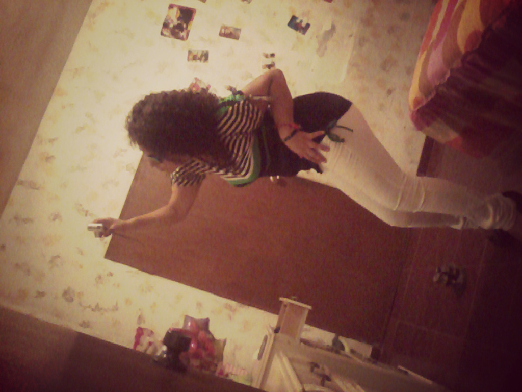 Eine Frau hält eine Kamera in der Hand und trägt ein schwarzes Oberteil und weiße Jeans, vor einem weißen Hintergrund und einer braunen Tür, mit einem Bett in der oberen rechten Ecke des Bildes.