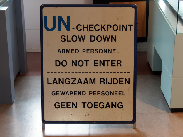 Ein Bodenaufkleber mit der Aufschrift "UN-Checkpoint Slow Down Armed Personnel - Do Not Enter" mit einer Glasplatte und Gegenständen rechts und einer Wand im Hintergrund.