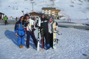 Vier Personen posieren für ein Foto mit Skating-Pads und Werkzeugen, mit Gebäuden und anderen Menschen im Hintergrund, vor einer schneebedeckten Berglandschaft.