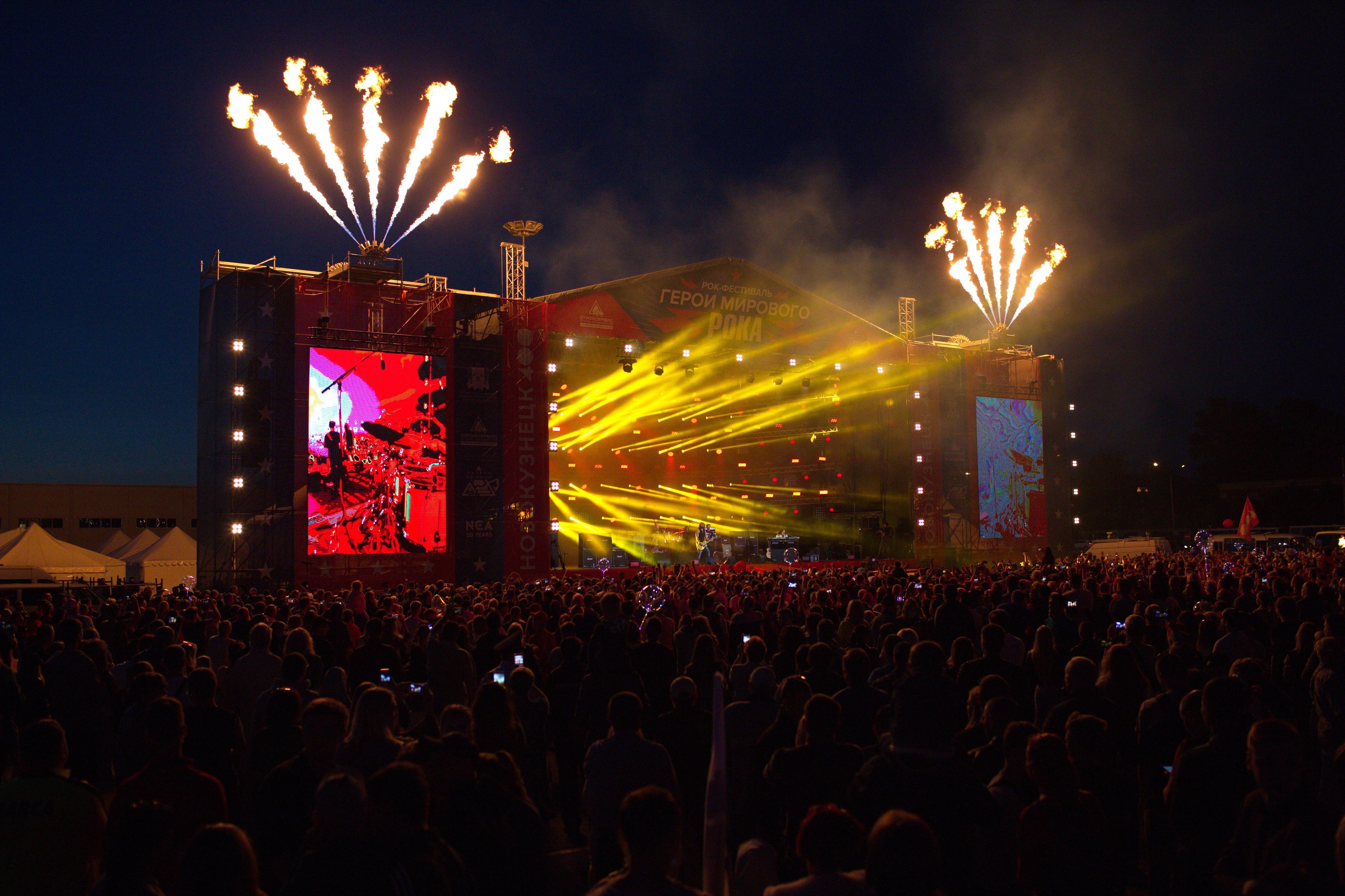 Eine große Menschenmenge auf einem Musikfestival schaut auf Performern auf einer hell erleuchteten Bühne, mit Zelten links und Feuerwerk in einem dunklen Himmel dahinter.