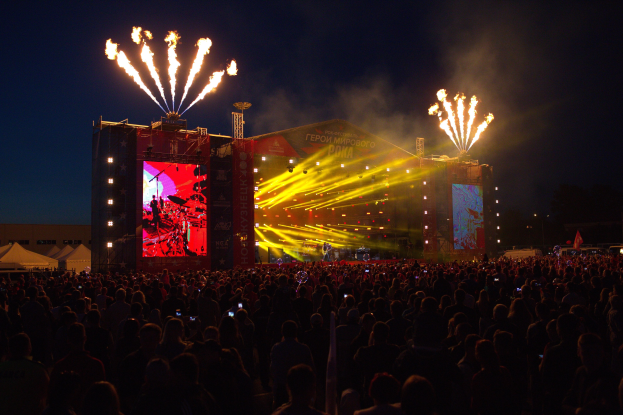 Eine große Menschenmenge auf einem Musikfestival schaut auf Performern auf einer hell erleuchteten Bühne, mit Zelten links und Feuerwerk in einem dunklen Himmel dahinter.