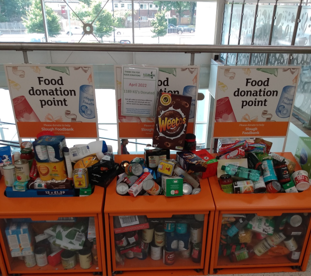 Orange Schübe mit Lebensmittelspenden gefülllt, darunter Flaschen, Dosen und Kisten, mit Informationsschildern, vor dem Hintergrund von Fahrzeugen, Gebäuden, Bäumen und Himmel durch Glasfenster sichtbar.