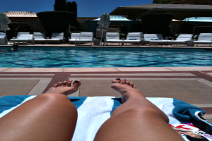 Frauen auf Poolbetten mit einem Schwimmbad und mehr Poolbetten mit Handtüchern im Hintergrund, umgeben von Büschen und Häusern.