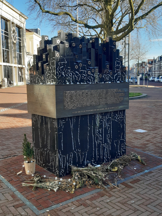 Denkmal in einem Stadtplatz für Holocaust-Opfer mit einem Baum davor, einem Topf mit einer Pflanze, verstreuten trockenen Blättern und Gebäuden, Fahrzeugen und Bäumen im Hintergrund.