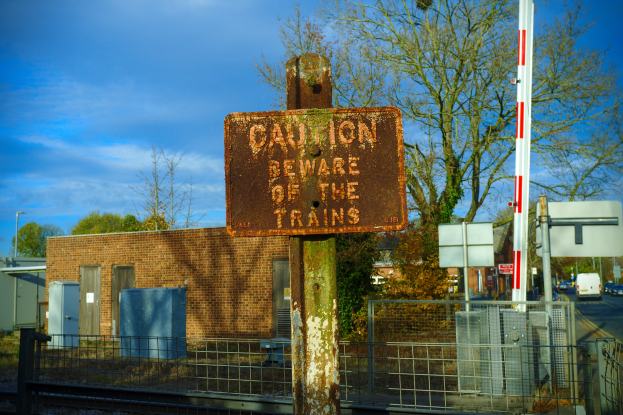 Vorsichtsschild an einem Bahngleiszaun mit Bäumen, Strommasten, einem Gebäude, Containern, Fahrzeugen und einem bewölkten Himmel.