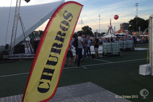 Eine lebhafte Karnevalsszene mit Zelten, einem zentralen Mast, Menschen, Bäumen, Strommasten, Straßenbahnen auf der Straße und einem Eisenständer links.
