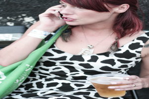 Eine Frau h√∂lt ein Glas Alkohol und ein Mobiltelefon, mit einer gr√ľnen Handtasche im Hintergrund.