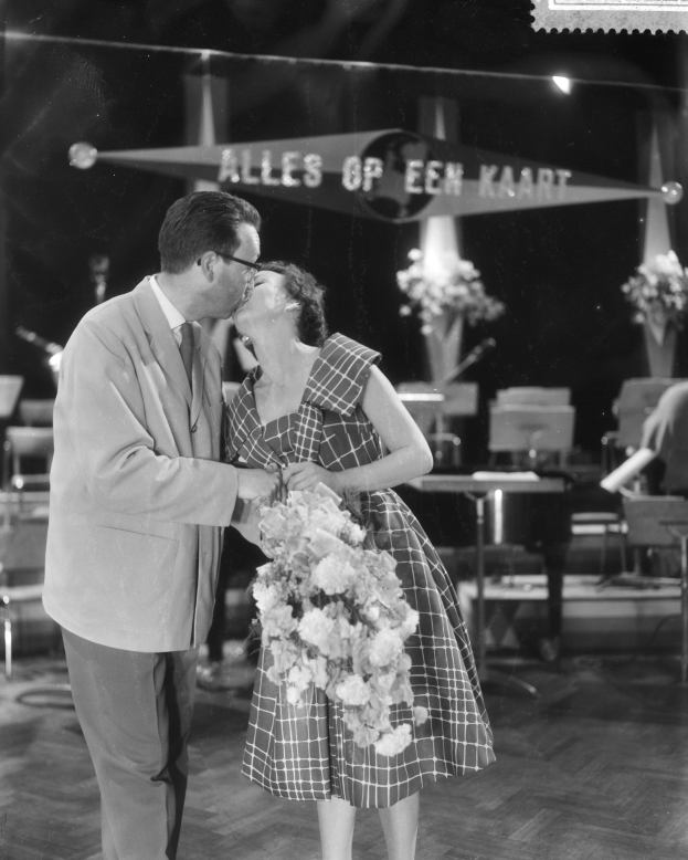Ein Mann und eine Frau umarmen und küssen sich auf einer Bühne, während die Frau einen Blumenstrauß hält und im Hintergrund Stühle und Blumenvasen zu sehen sind.