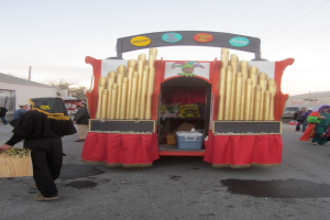 Ein geschmückter Lkw mit Gegenständen darin, zwischen Scheunen geparkt, mit Menschen in der Nähe, Autos auf beiden Seiten, Bäume davor und Kabel darüber.