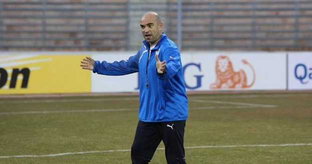Ein Fußballtrainer in einer blauen Jacke steht auf einem Feld während des Trainings, umgeben von Tafeln mit Text und Logos, und sieht entschlossen aus.
