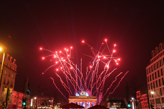 Eine belebte Stadtstraße an einem Silvesterabend in Berlin mit Gebäuden, Bäumen, Laternenmasten, Verkehrsampeln, Schildern, Zelten, Menschen und einem prächtigen Feuerwerk am Himmel.