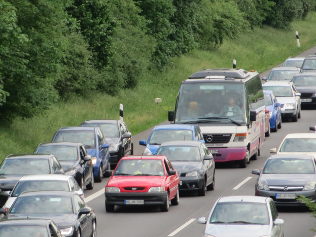 Ein Stau auf einer Autobahn mit vielen Autos und einem Van, Menschen in den Fahrzeugen und Bäume und Gras im Hintergrund.