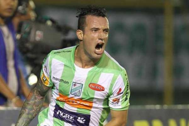 Ein Fußballspieler in grüner und weißer Uniform mit Tattoos auf dem Arm feiert nach einem Tor, mit unscharfen Figuren und einer Tafel im Hintergrund.