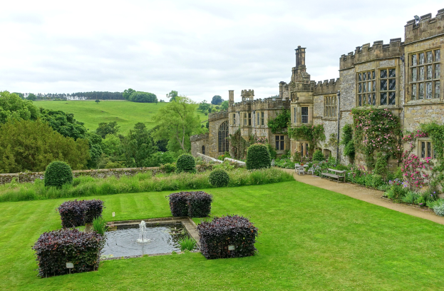 Gro├če Geb├Ąude mit einer zentralen Font├Ąne, umgeben von Grün, einem Weg und einer Bank unter einem teilweise bew├Âlktem Himmel, wahrscheinlich Stourhead Castle.