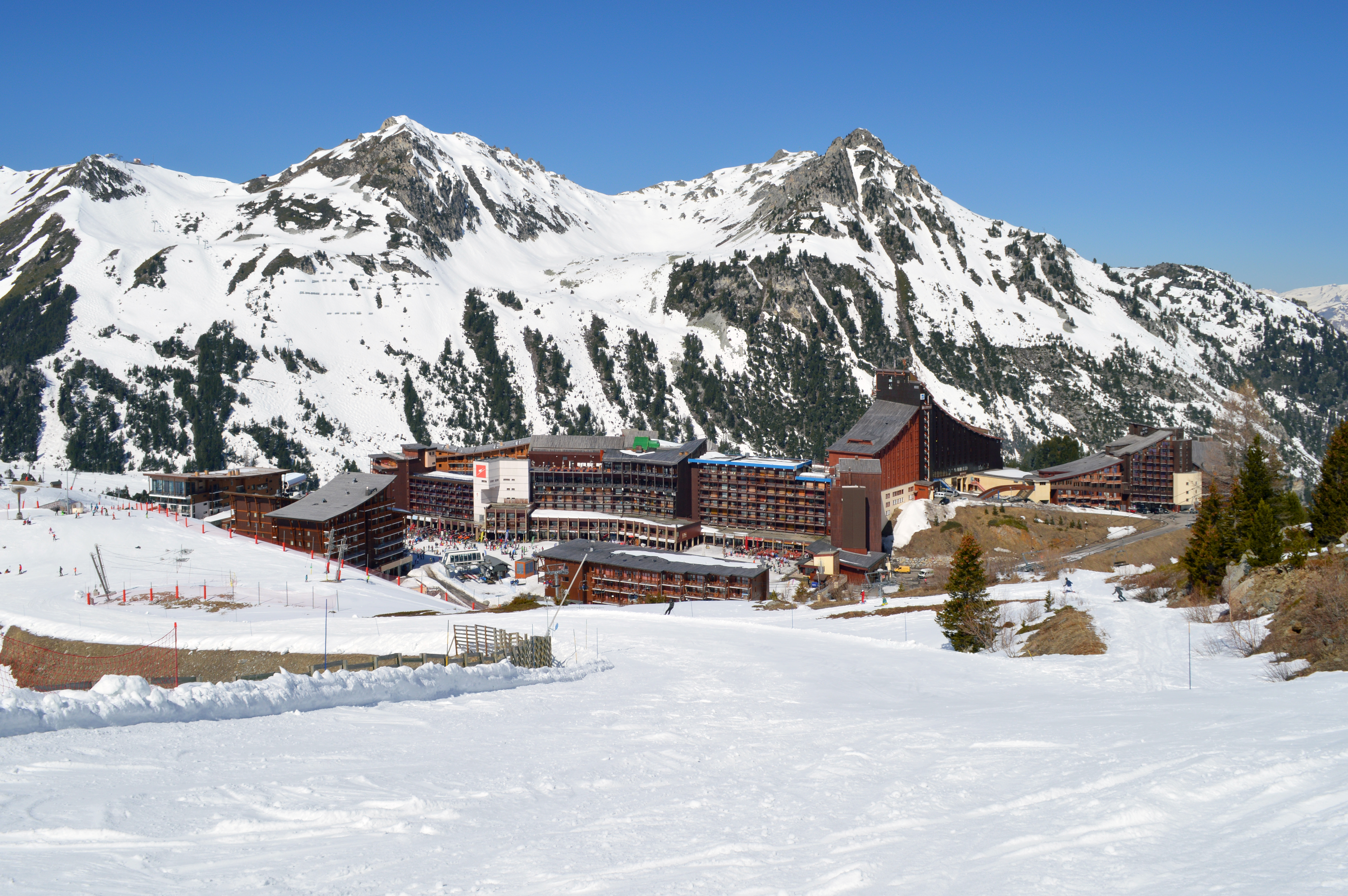 Ein Skigebiet mit schneebedeckten Bergen im Hintergrund, weißer Boden, verstreute Bäume und Gebäude, unter einem strahlend blauen Himmel.
