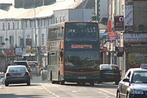 Eine Straße mit Autos und einem Bus im Vordergrund, im Hintergrund Gebäude mit Wänden, Fenstern, Tellern und Dächern sowie Plakate und Banner an den Wänden und ein Laternenmast.