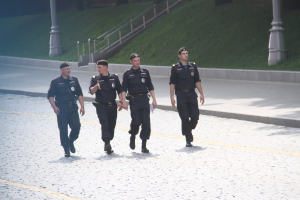 Drei Polizisten in Uniform und Mützen, die eine Straße mit Geländern, Treppen und Pfählen im Hintergrund entlanggehen.