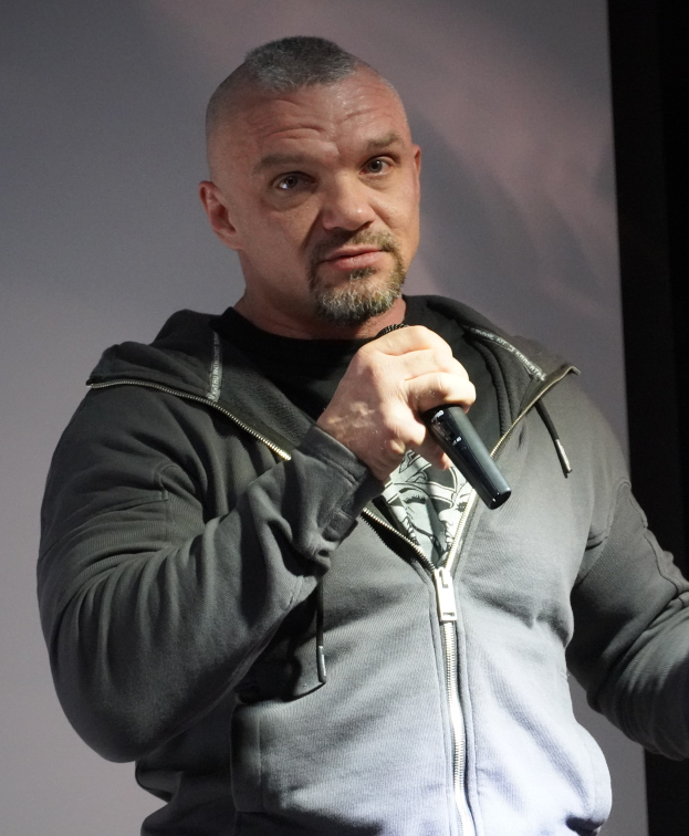 Ein Mann in einer schwarzen Kapuzenjacke und Anzug, der ein weißes Hemd und zurückgekämmtes Haar trägt, hält ein Mikrofon mit einem selbstbewussten Ausdruck vor einer Wand.