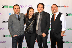 Eine Gruppe strahlender Menschen posiert vor einer Kamera mit einem Sponsorenbanner im Hintergrund.