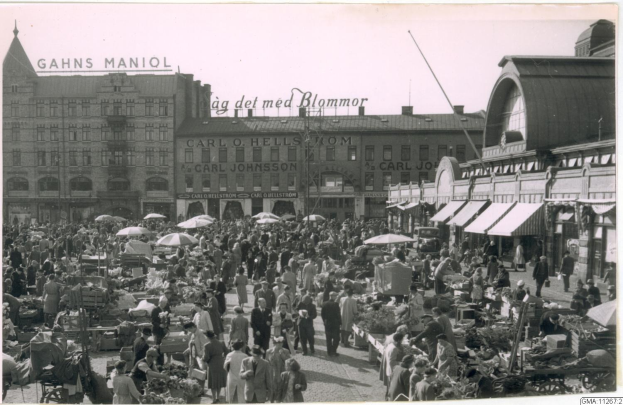 Schwarz-weiß-Foto eines belebten Berliner Markts mit Menschen, Gemüsekarren und Gebäuden im Hintergrund.