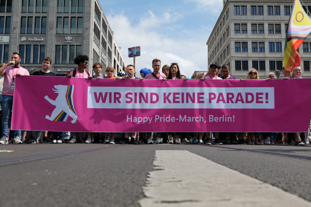 Eine Gruppe von Menschen bei einer Pride-Veranstaltung in Berlin, die eine pinkfarbene Fahne mit der Aufschrift 'Happy Pride March Berlin' schwenken, wobei einige eine Kappe und eine Schutzbrille tragen und einer eine Fahne hält, vor dem Hintergrund von Gebäuden, einem Schild und einem bewölkten Himmel.