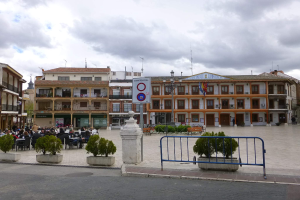 Ein belebter Stadtplatz mit Menschen, die sitzen und stehen, Topfpflanzen, Metallabsperrungen, ein Schild an einem Pfahl, Straßenlaternen mit Fahnen, Gebäude mit Fenstern und eine bewölkte Himmel.