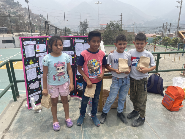 Gruppe von Kindern auf einem Basketballfeld mit Papiertüten in den Händen, mit Tüten und Gegenständen rechts und einer Tafel mit Text links, vor einem Hintergrund aus Zaun, Pfählen, Straßenlaternen, Strommasten, Bäumen, Gebäuden, Hügeln und Himmel.