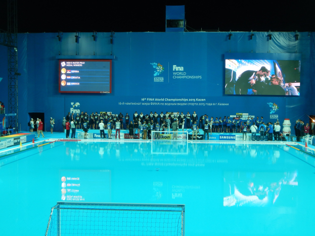 Gruppe von Menschen, die um ein Hallenbad mit Netz am Boden stehen, mit einer Wand im Hintergrund, die "FINA World Championships 2015 Kazan"-Text und -Logos, Träger und Lampen anzeigt.