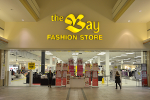 Außenansicht eines Bay Fashion Stores in einem Einkaufszentrum mit Menschen, Texttafeln an den Wänden, Säulen, Deckenleuchten und Hintergrundauslagen.