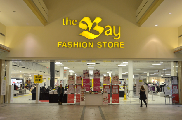 Außenansicht eines Bay Fashion Stores in einem Einkaufszentrum mit Menschen, Texttafeln an den Wänden, Säulen, Deckenleuchten und Hintergrundauslagen.