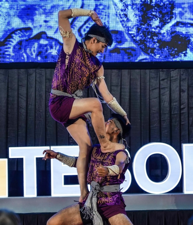 Zwei Akrobaten performen auf der Bühne, der Mann hält die Frau in seinen Armen, während beide purple Kleider tragen, mit einem Publikum unten und einem schwarzen Vorhang und Bildschirm im Hintergrund.