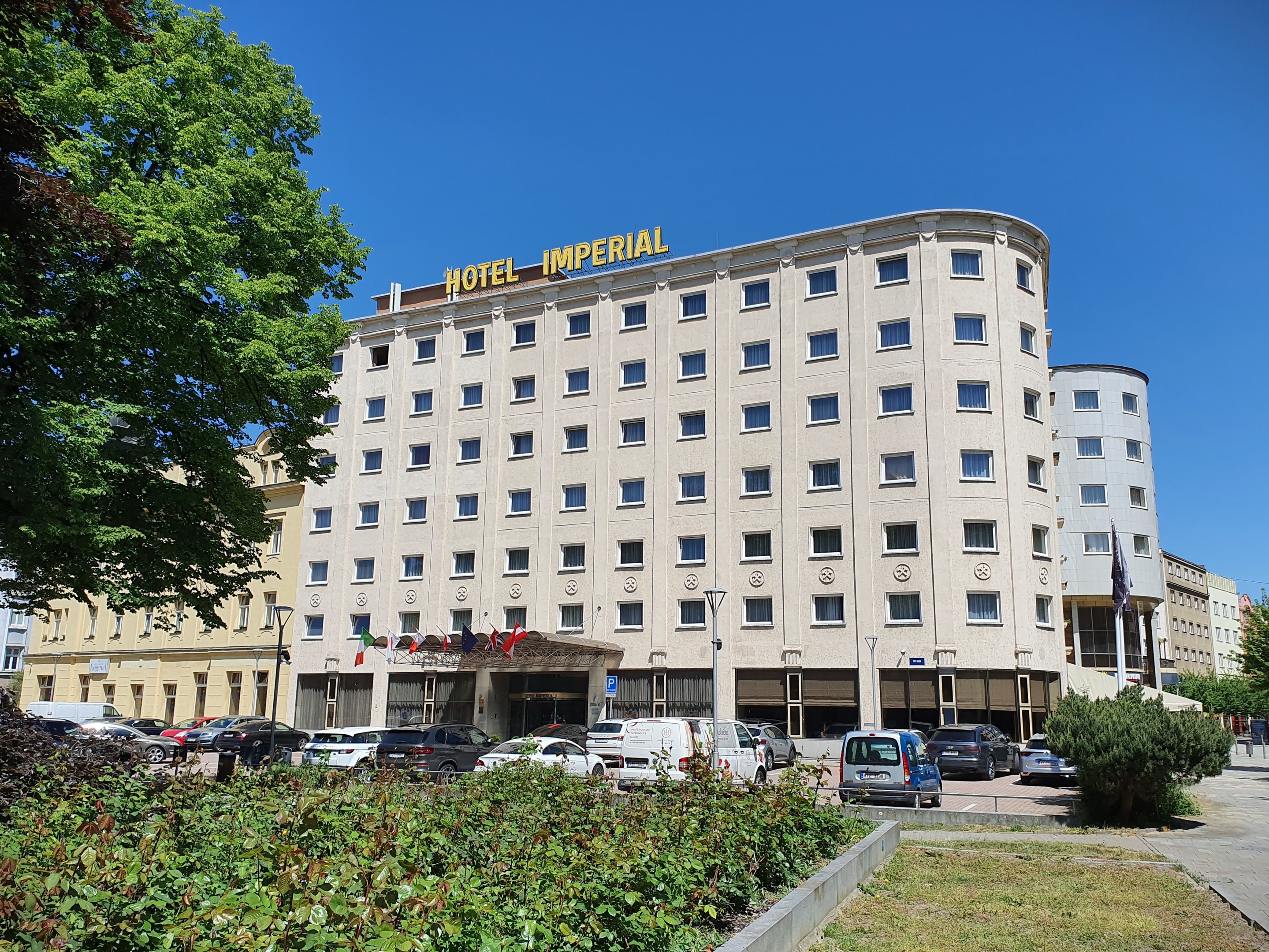 Das Hotel Imperial in Berlin, ein großes Gebäude mit Fenstern, umgeben von Grün, geparkten Autos, Straßenmasten mit Flaggen, unter einem bewölkten Himmel.