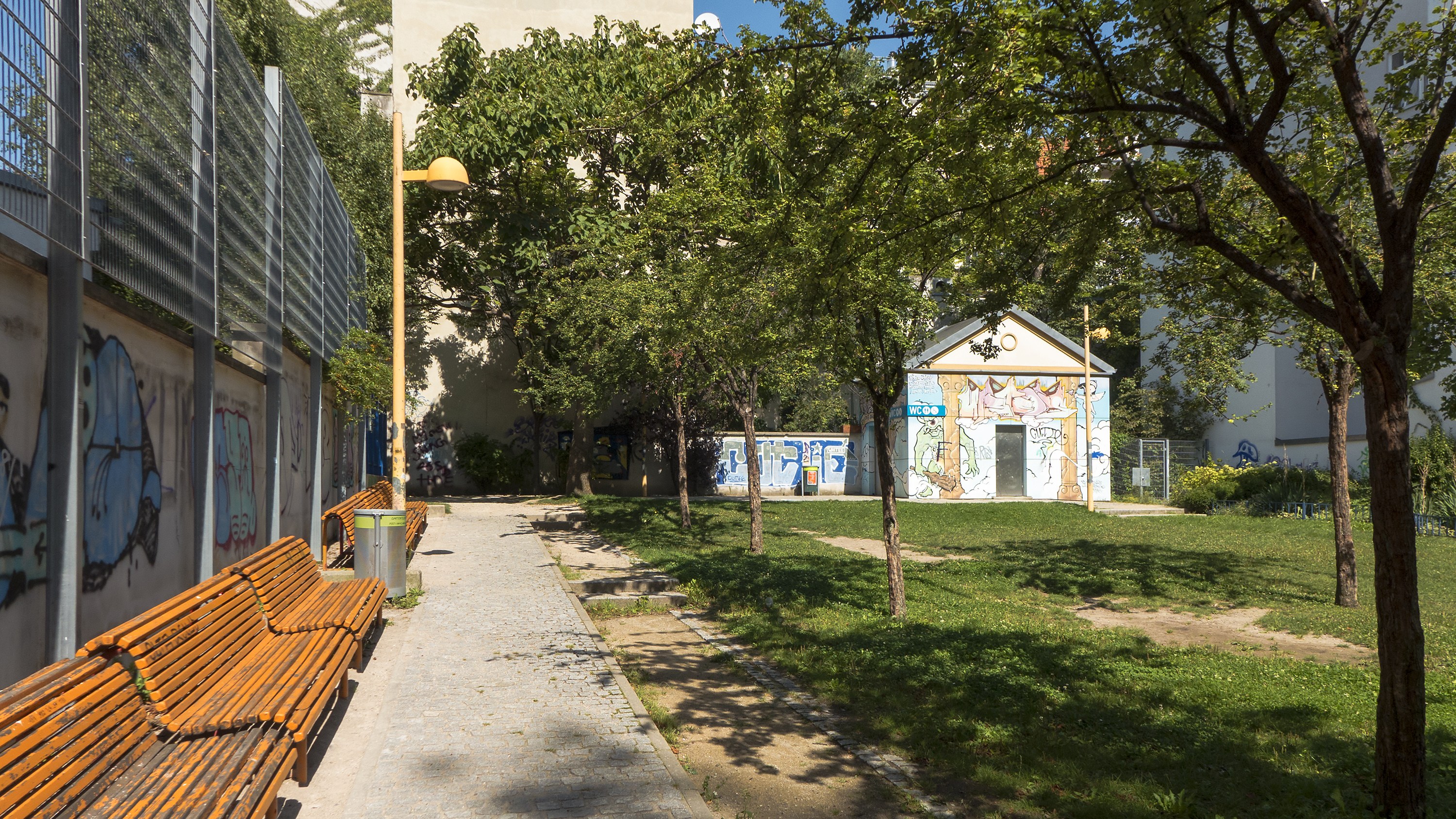 Ein Park mit Bänken, Graffiti an einem Gebäude, Bäume, Gras, ein Mülleimer, eine Straßenlaterne, ein Zaun, entfernte Gebäude und einen klaren blauen Himmel.