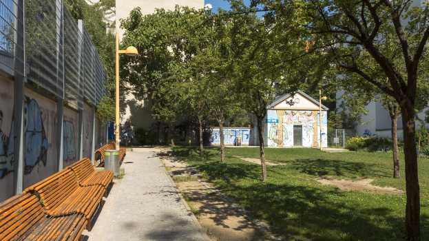 Ein Park mit Bänken, Graffiti an einem Gebäude, Bäume, Gras, ein Mülleimer, eine Straßenlaterne, ein Zaun, entfernte Gebäude und einen klaren blauen Himmel.