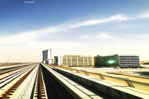 Eisenbahnschienen in der Mitte, mit Gebäuden und Straßen auf der rechten Seite und einem Himmel mit Wolken im Hintergrund.