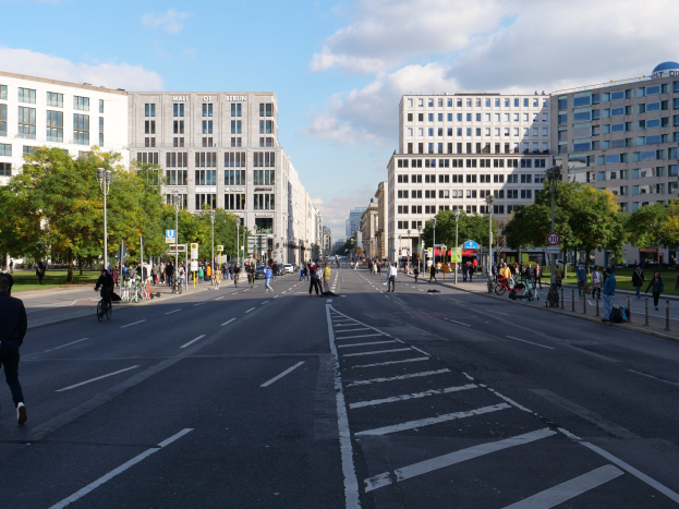 Eine belebte Stadtstraße in Berlin, Deutschland, mit Fußgängern und Radfahrern auf der Straße, hohen Gebäuden mit Fenstern, Bäumen, Laternenmasten und Schildern, unter einem bewölkten Himmel.