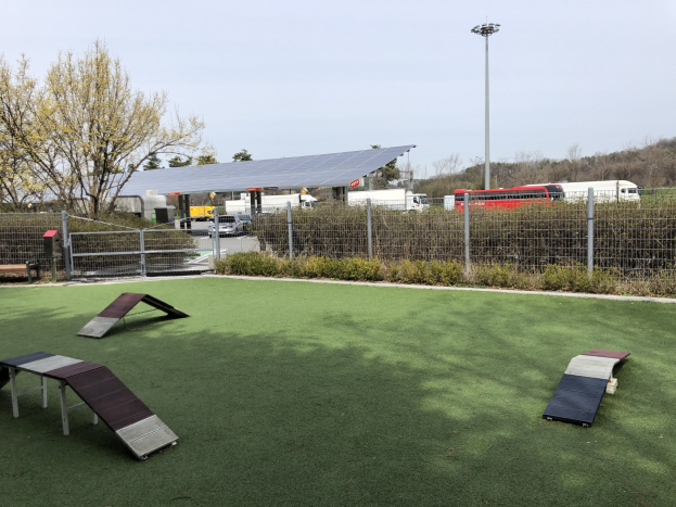 Hundepark mit Kunstgras, Rampen, einem Zaun, Pflanzen, Bäumen, Fahrzeugen, Häusern, Pfählen und einer Laterne.