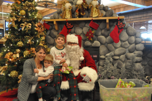 Eine Frau, Kinder und ein Mann als Santa Claus mit Brille und Handschuhen stehen neben einem geschm√ľckten Weihnachtsbaum, einer Wand mit Steinen, Spielzeugen, Dekorationen und einem Korb mit Gegenst√§nden, mit Lichtern im Hintergrund.