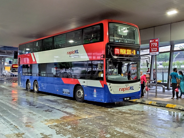 Ein roter und blauer Doppeldeckerbus steht auf einem Parkplatz mit Menschen, die herumlaufen, Taschen tragen, von einem Metallzaun umgeben, einem Schild, einem Dach mit Deckenleuchten und Säulen.