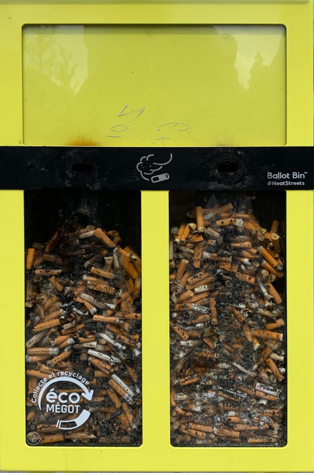 Gelber Mülltonne mit Zigarettenstummeln auf einem Gehweg, mit Text auf der Tonne.