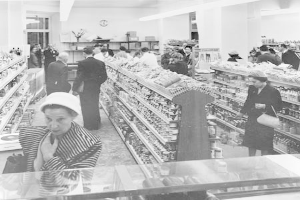 Ein Schwarz-Weiß-Foto von Menschen, die in einem Supermarkt einkaufen, mit Taschen und Lebensmittelregalen im Bild, einer Uhr an der Wand und Deckenlampen.