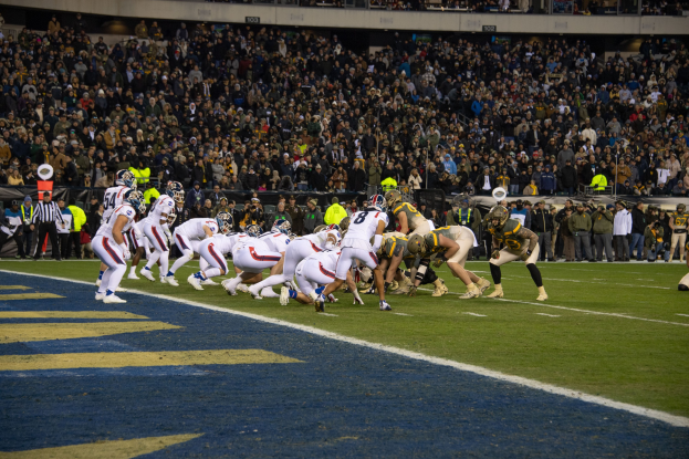 Ein Fußballspiel im Gange auf einem Stadionfeld mit Spielern in Sportbekleidung und Helmen auf dem Boden, umgeben von Zuschauern und Stadionstrukturen.