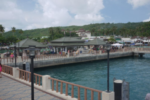 Ein breiter Fluss mit einer ├╝ber ihm stehenden Br├╝cke, Menschen unter einem Zelt auf der Br├╝cke, ein Berg mit B├Ąumen und Geb├Ąuden im Hintergrund.