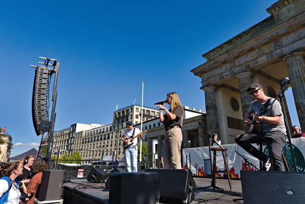 Eine Gruppe von Menschen, die auf einer Bühne Musik spielen, mit dem Brandenburger Tor im Hintergrund, begleitet von Equipment und umgeben von städtischer Szenerie.