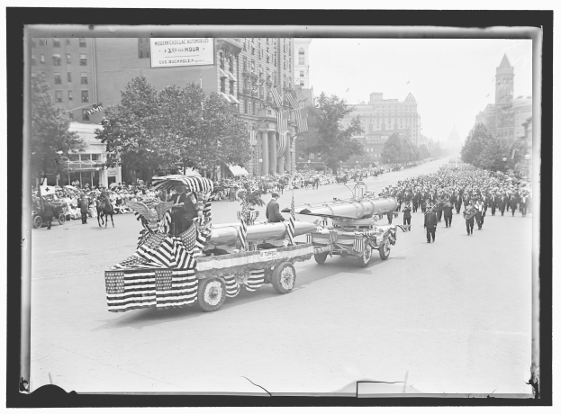 Schwarzes und weißes Foto eines Umzugs mit einem Löschfahrzeug in der Mitte, umgeben von Menschen, einige halten Fahnen, mit Bäumen, Gebäuden und einer Tafel mit Text im Hintergrund.